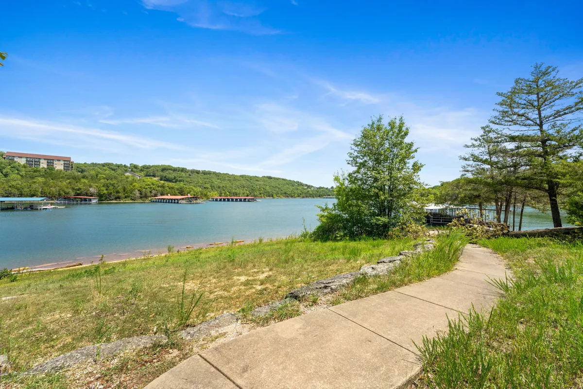 A peaceful scene featuring a lakeside pathway surrounded by trees, with the calm waters of Table Rock Lake and distant hills in view. Boathouses dot the shoreline, and the natural setting invites leisurely walks, fishing, and exploring the outdoors.