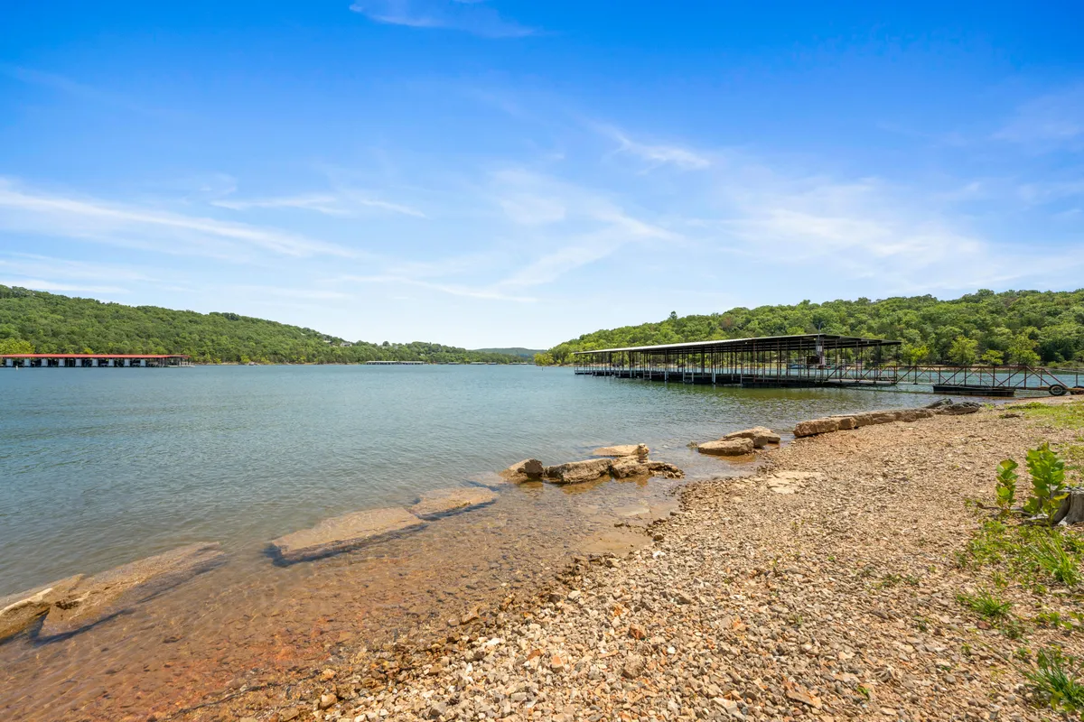 A scenic lake view with calm water bordered by a rocky shoreline, distant green hills, and a boathouse structure. The setting is inviting for fishing, boating, and water sports, with an atmosphere of serene outdoor adventure.