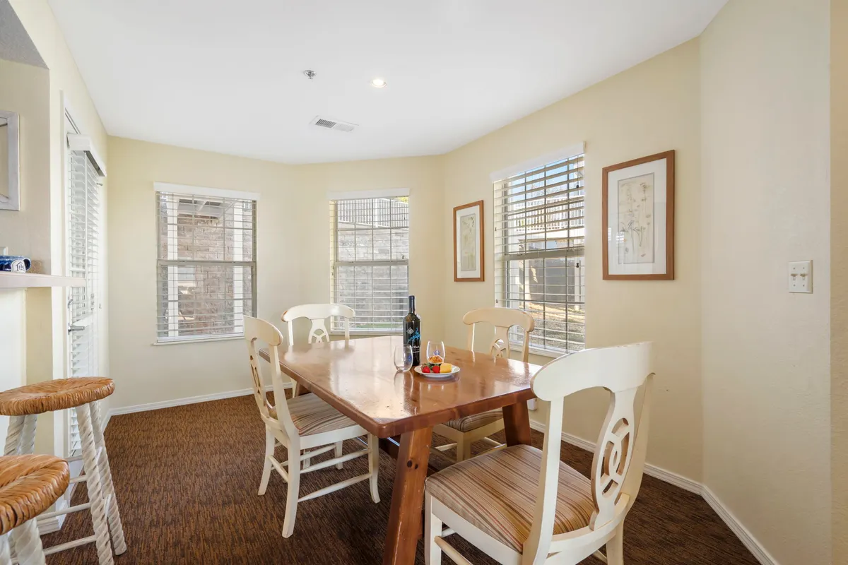 A welcoming dining area centered around a wooden table, with a bottle of wine and decorative fruit bowl serving as focal points. Framed artwork and large windows contribute natural light and a cheerful, homelike ambiance. The table is set for meals and socializing, suggesting both casual morning coffees and more formal gatherings.