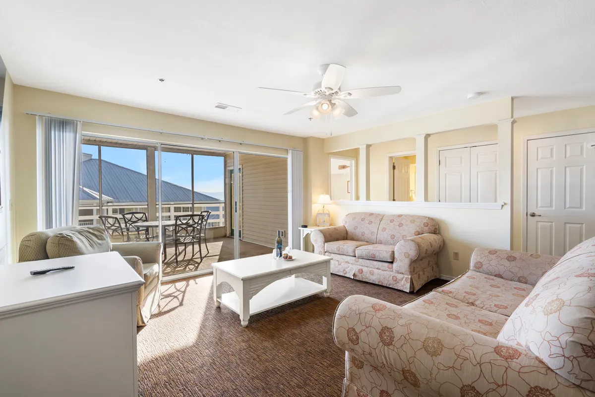 This image captures the heart of the living area, with plush sofas, a coffee table, and expansive windows that bathe the space in natural light. An inviting outdoor seating area is visible through the windows, providing a glimpse of the seamless connection between indoors and the private deck outside.