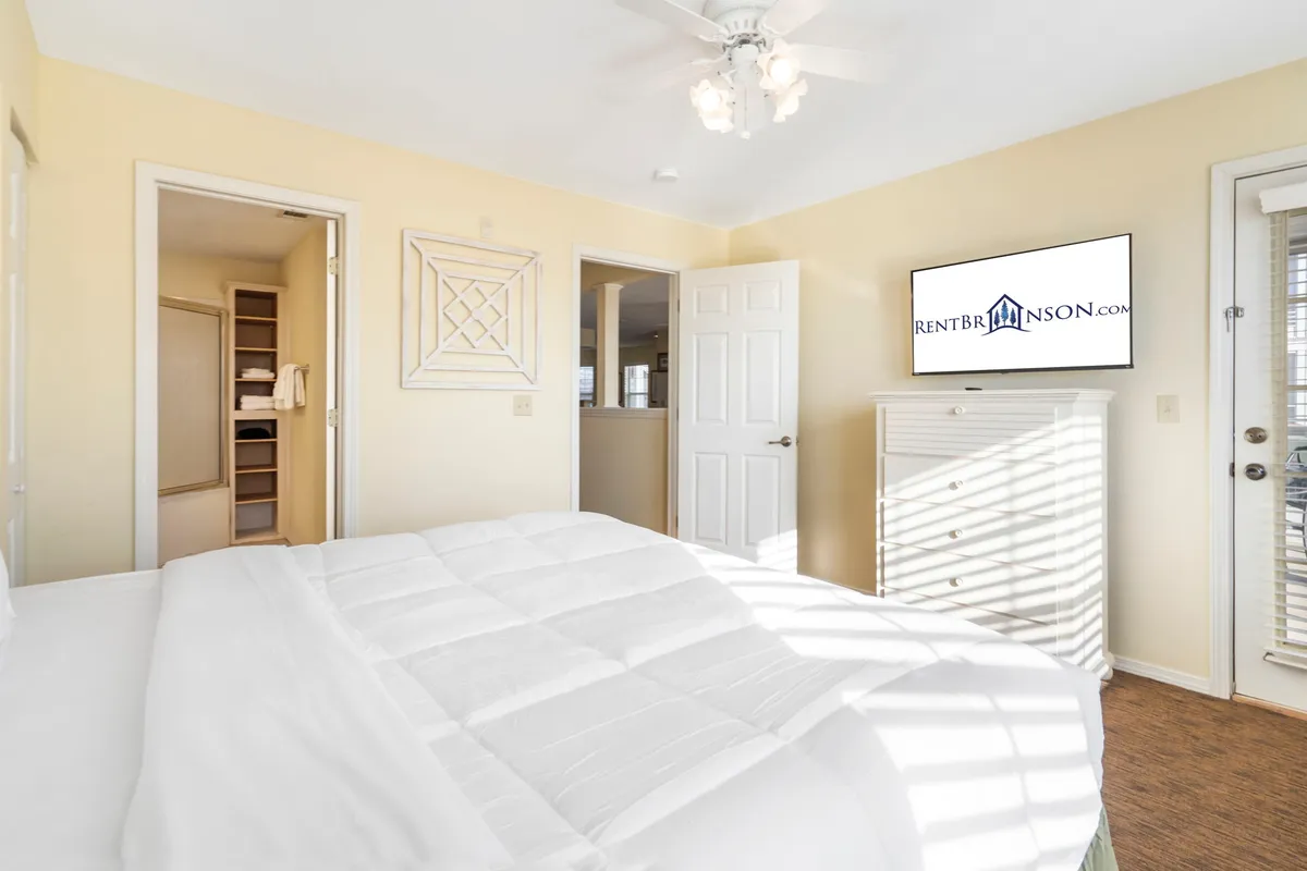A spacious bedroom with a large king-sized bed, soft bedding, a wall-mounted television, and doors leading to another space. The room is styled for comfort, hinting at restful nights and cozy relaxation. Ambient lighting and neutral tones create a tranquil atmosphere.