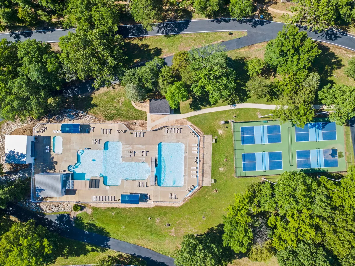 An expansive view of the resort's outdoor amenities, including sparkling swimming pools, tennis and pickleball courts, and lush surrounding greenery. The scene captures both relaxation and active recreation, with people swimming, sunbathing, and playing sports under the cheerful sky.