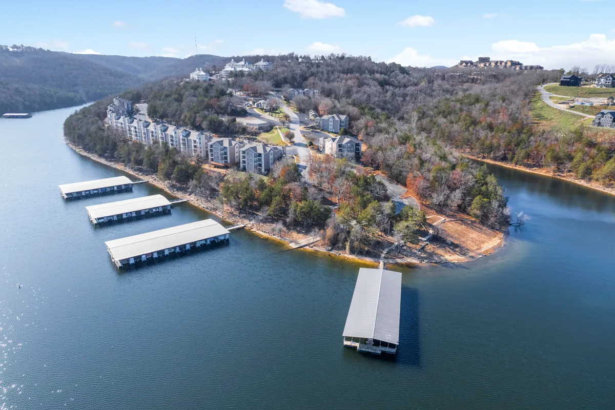 A tranquil lakeside scene shows Table Rock Lake, with a dock stretching out over calm waters, rolling hills in the background, and residential buildings nearby. The area beckons for swimming, fishing, boating, or simply relaxing by the water's edge.