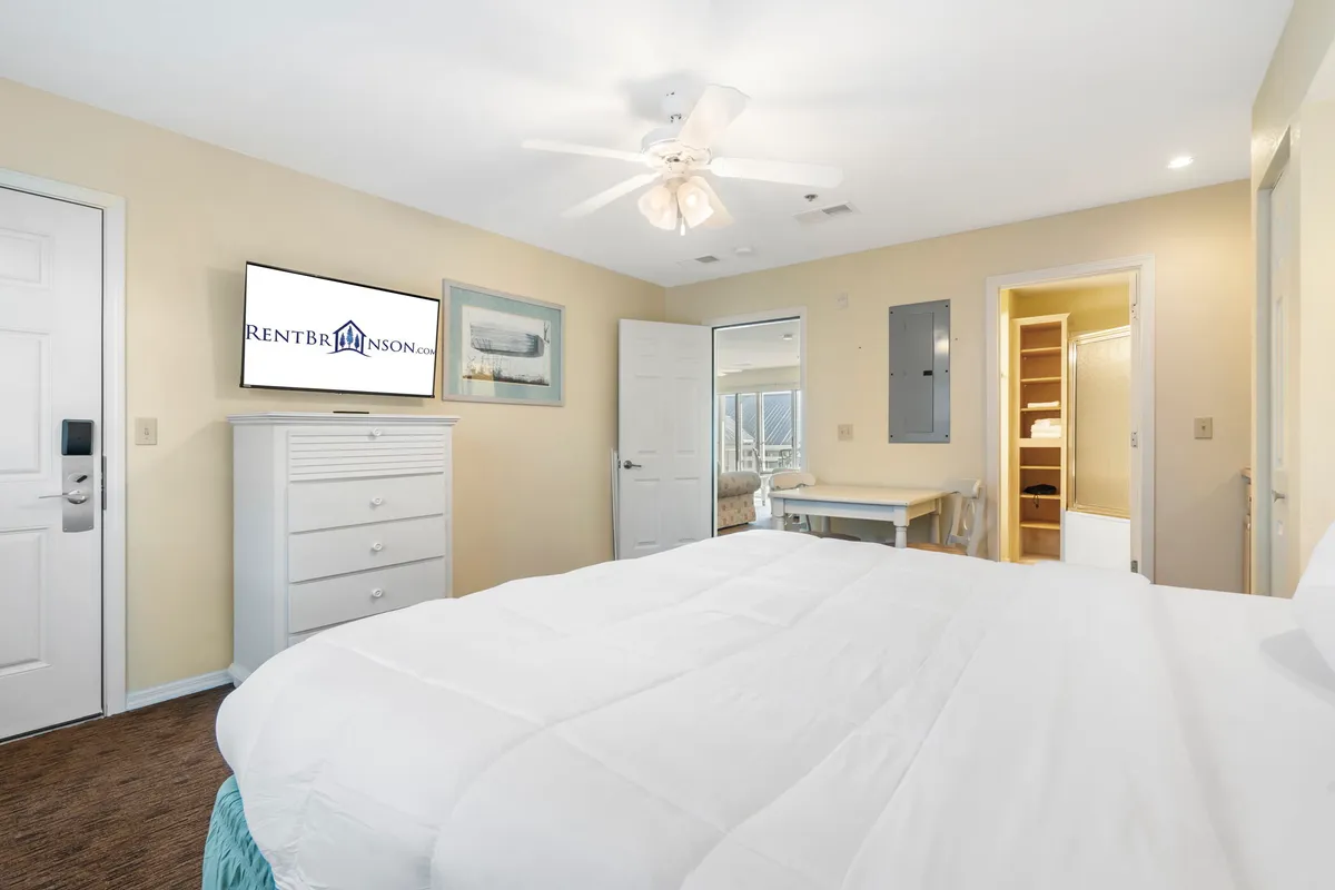 A second bedroom featuring a king bed with white bedding, a dresser with a television, open closet, and tasteful wall art. Sunlight softly illuminates the room, offering a cozy and inviting environment ideal for relaxation and unwinding.